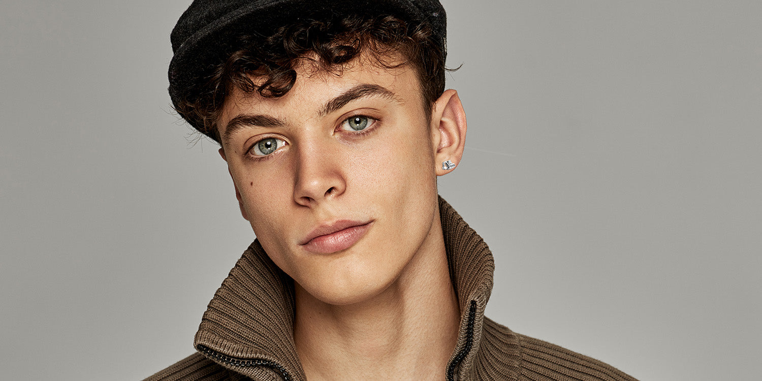 unger Mann mit lockigen Haaren und schwarzer Mütze in braunem Strickpullover  trägt silbernen Ohrstecker mit Bagger-Motiv